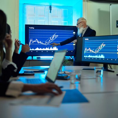 Bearded male worker showing graphs on monitor while speaking against anonymous colleagues at table in office